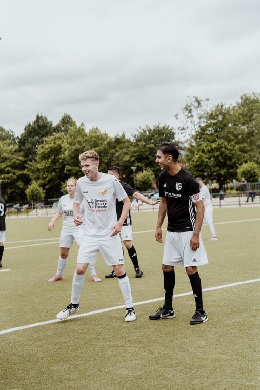 Zwei Spieler lachen auf dem Platz miteinander, einer im weißen, einer im schwarzen Trikot