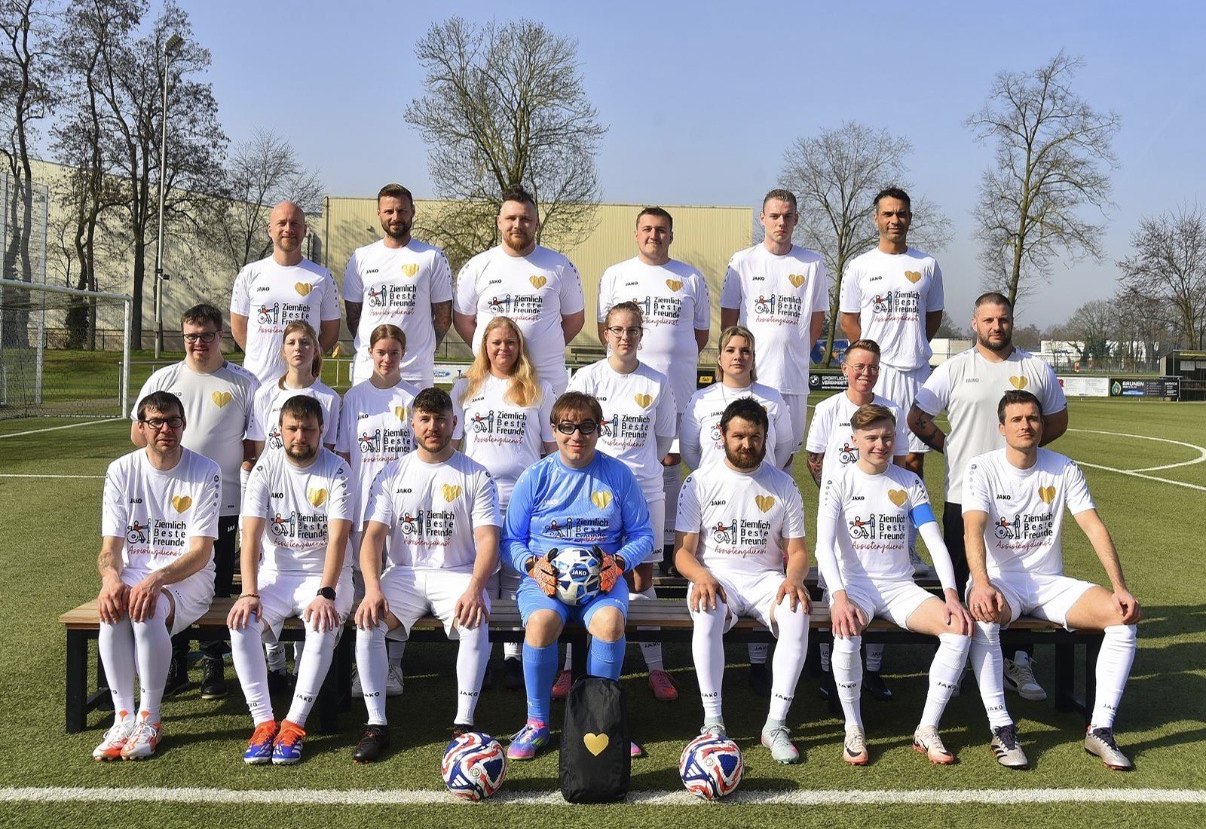 Die Inklusionsmannschaft in drei Reihen auf dem Sportplatz, in weißen Trikots mit dem goldenen Herz, in der Mitte der Torwart im blauen Trikot mit dem Ball auf dem Schoß